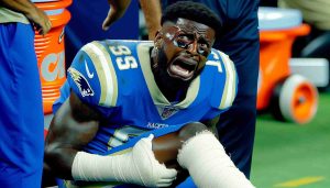 An ultra high definition photo that captures the candid moment of a star running back, who is of African descent, visibly in pain during a football match. He is shown sitting on the sidelines, his leg bandaged, as he grimaces in pain. His future participation in upcoming games appears uncertain, a fact reflected in his incredulous and distressed expressions.